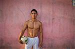 Portrait of a shirtless young man holding a soccer ball while standing against a wall.