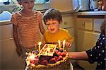 Young boy blowing out his birthday candles as his smiling mother holds the birthday cake in their kitchen.