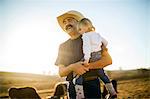 Cowboy rancher holding his toddler daughter in his arms while they are out on the ranch.