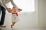 Sweet smiling baby girl in a floral dress takes her first steps assisted by her daddy.