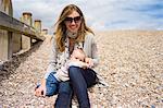 Mother with baby girl sitting on shingle beach looking at camera smiling