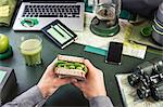 Mans hand's holding sandwich at table with hiking equipment and laptop