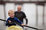 Young boy sticks his tongue out in concentration as he tries to row a kayak while his father helps to push it along in the waters of a foggy harbour.