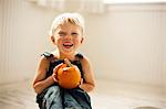 Young boy holds a small Jack O'Lantern on his knees as he sits on a floor and poses for a portrait.