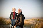 Father and his small son smile as they pose for a portrait while kneeling and standing on a grassy sand dune.