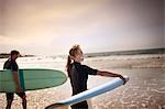 Young woman grins and looks over her shoulder as she carries a surfboard into the sea.