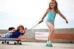Young girl pulling her brother on a makeshift wagon,  made from a boogie board on top of a skateboard.