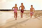 Portrait of three smiling children running together along a sandy beach wearing bathing suits.