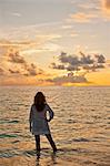 Young woman standing ankle deep in the tropical ocean while watching the beautiful sunset.