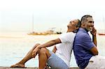 Happy mature couple sitting back to back by the beach.