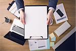 Businessman working at office desk and showing a clipboard with a blank document, computers and stationery on background, top view