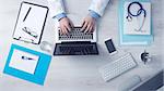 Doctor sitting at office desk and working on his laptop with medical equipment all around, top view