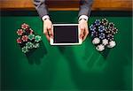 Male player holding a digital tablet and playing with stacks of chips all around on a green table, top view