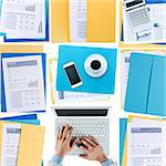 Businessman working at desktop on a laptop with financial reports, paperwork and files, top view, unrecognizable person