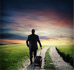 Businessman with luggage walk on road in the countryside with green grass at sunset