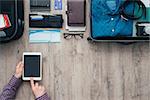 Man packing for a trip, he is using a digital touch screen tablet and booking flights online using a mobile app, flat lay banner