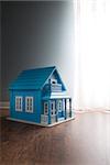 Blue wooden model house next to a window with curtain on wooden floor.