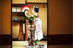 A woman dressed in the traditional geisha style, wearing a kimono and obi, with an elaborate hairstyle and floral hair clips, with white face makeup with bright red lips and dark eyes. Standing in a classic pose with fan raised, side view.