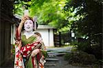 A woman dressed in the traditional geisha style, wearing a kimono and obi, with an elaborate hairstyle and floral hair clips, with white face makeup with bright red lips and dark eyes taking a selfie.