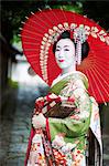 A woman dressed in the traditional geisha style, wearing a kimono and obi, with an elaborate hairstyle and floral hair clips, with white face makeup with bright red lips and dark eyes holding a red paper parasol.