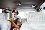 A woman dressed in the traditional geisha style, wearing a kimono with an elaborate hairstyle and floral hair clips, with white face makeup with bright red lips and dark eyes in a car using a smart phone