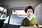 A woman dressed in the traditional geisha style, wearing a kimono with an elaborate hairstyle and floral hair clips, with white face makeup in a taxi, using a smart phone.