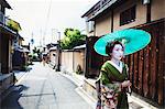 A woman dressed in the traditional geisha style, wearing a kimono and obi, with an elaborate hairstyle and floral hair clips, with white face makeup with bright red lips and dark eyes holding a paper parasol walking along a street.