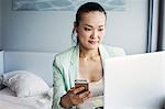 A business woman preparing for work, sitting on a bed using a laptop and holding her smart phone.