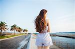 Back view of young woman with long red hair standing in open space by a road on the coast.