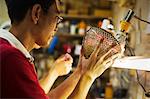 A craftsman in a glass maker's workshop holding a vase with a delicate geometric pattern up against a light.