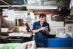 A traditional fresh fish market in Tokyo. A man in a blue apron standing behind the counter of his stall, using a smart phone covered with protective plastic.