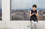 A woman seated by a window with a view over a large city with her back to the view, using her smart phone.