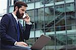 Businessman talking on mobile phone while using laptop outside side