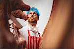 Butcher looking at the red meat hanging in storage room at butchers shop