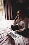 Pregnant woman talking on mobile phone while using laptop in bedroom at home