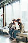 Male adult hipster twins sitting whilst working on laptop in office