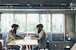 Male adult hipster twins sitting face to face at desk in office