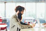 Young male hipster with curly hair and beard using digital tablet at desk