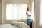 Unhappy mature woman in her pajamas stands with her arms crossed in front of a bedroom window as she poses for a portrait.