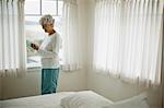 Mature woman in her pajamas smiles as she looks down at a cell phone as she stands in front of a bedroom window.