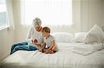 Mature woman in her pajamas sits on a bed with her young grandson and shows him a cell phone.