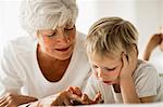 Mature woman and grandson lie on their fronts on a bed side by side while looking down.