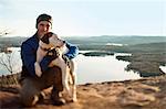 Man embraces his dog as he poses for a portrait above a scenic view of a lake,  islands and woodlands.