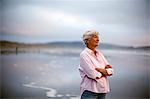 Content mature woman with her arms crossed stands on a beach and looks out at the sea.