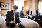 Businessman sits on the edge of a bed in a hotel room during a business trip and writes in a folder while his three female colleagues work together in the background.