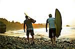 Two men walk along a beach carrying a surfboard each as the sun rises ahead of them.