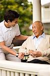Male nurse laughs and talks with a senior man in a wheelchair as they sit on a porch.