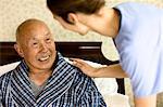 Senior man sitting in bed smiles up at a female nurse as she leans over to put a reassuring hand on his shoulder.