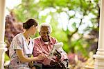 Female nurse laughs and talks with a senior man as they sit on a porch and look at a newspaper.