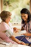 Female nurse uses a blood pressure cuff and blood pressure monitor on a senior woman who sits on a bed.